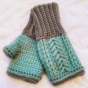 May include: A pair of crocheted fingerless gloves in a teal and grey colour scheme. The gloves feature a cable knit pattern on the back of the hand and a simple stitch on the palm.