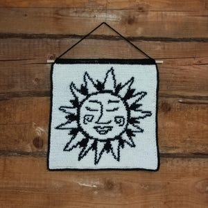 May include: A white crocheted wall hanging with a black crocheted sun with a smiling face.