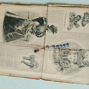 Könnte beinhalten: Vintage Seiten mit Modeillustrationen, mit Bindfaden gebunden und mit blauen floralen Akzenten verziert. Die Seiten zeigen detaillierte Zeichnungen von Damenbekleidung und Accessoires, die ein historisches Modethema aufweisen.