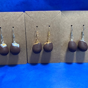 May include: Three pairs of earrings with brown and gold coconut-shaped charms hanging from silver and gold hooks. The earrings are displayed on brown card stock.