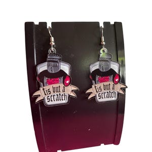 May include: Dangle earrings with a cartoon knight design. The knight wears a black helmet and armour, with a red heart detail. A banner below reads "tis but a scratch". The earrings hang from silver-coloured hooks.