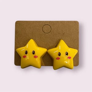 May include: A pair of yellow star-shaped earrings with pink cheeks and black eyes.