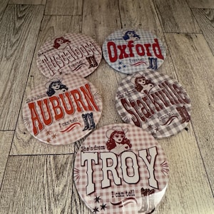 May include: Five circular coasters featuring a gingham pattern background. Each coaster displays a graphic of a woman in a cowboy hat, the text 'She's from' and 'I can tell', a pair of cowboy boots, and the city names Tuscaloosa, Oxford, Auburn, Starkville, or Troy.