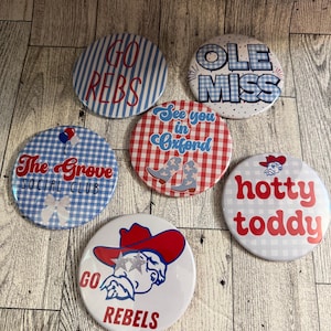 May include: Six circular pinback buttons featuring various designs in red, white, and blue. Text includes GO REBS, OLE MISS, The Grove SOCIAL CLUB, See you in Oxford, hotty toddy, and GO REBELS, with graphics of cowboy boots, a bow, and a mascot.