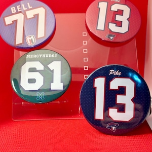 May include: Four round buttons with sports-themed designs. One button has "BELL 77" in white on a purple background. Two buttons display "13" in white, one on red and one on blue. The fourth button has "MERCYHRUST 61" in white on a green background.