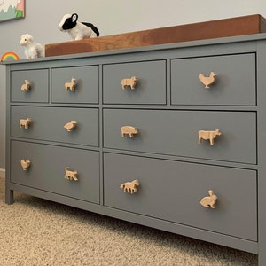 May include: A gray dresser with six drawers and wooden animal drawer pulls. The dresser has a light wood top and is in a nursery with a pink and white blanket on a rocking chair, a rainbow wall hanging, and animal wall art.