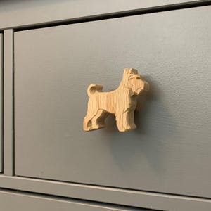 May include: A wooden dog-shaped cabinet knob fixed to a grey drawer. The knob is made from light-coloured wood, showing the grain, and is carved in detail. The dog-shaped knob provides a decorative element to the furniture.