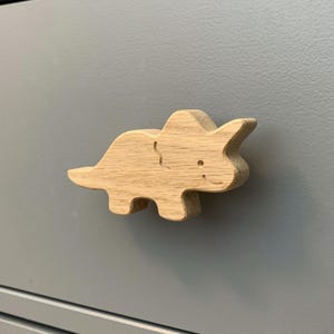 May include: A wooden dinosaur-shaped cabinet knob. The knob is light brown, with a detailed carving of a triceratops. The dinosaur has three horns, a frill, and a smiling face. The knob is mounted on a gray cabinet door.