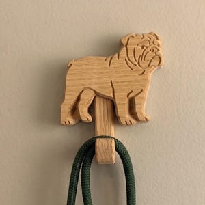 May include: Wooden bulldog-shaped wall hook. The hook is made of light-colored wood and features a detailed carving of a bulldog. A dark green rope is hanging from the hook, which is mounted on a neutral-colored wall.