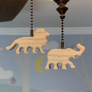 May include: Two wooden animal-shaped ceiling fan pull chains. One is a lion, the other an elephant. Both are made of light-colored wood and hang from a dark brown ceiling fan fixture. The background is a light blue wall with a moon.