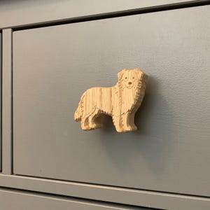 May include: A wooden dog-shaped cabinet knob. The knob is carved from light-coloured wood and features a detailed depiction of a dog. The knob is mounted on a grey cabinet drawer, providing a decorative and functional element.