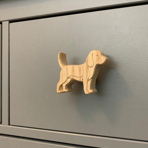 May include: A light brown, wooden dog-shaped cabinet knob. The knob is layered and detailed, depicting a dog's form. It is attached to a grey cabinet door, providing a decorative and practical feature for furniture.