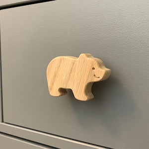 May include: A wooden bear-shaped cabinet knob. The knob is made of light-colored wood and has a simple, cartoon-like design with a smiling face. It is mounted on a gray cabinet door. The knob is approximately 3 inches long.
