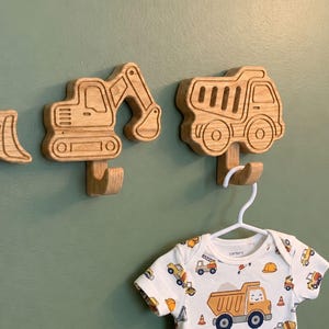 May include: Wooden wall hooks shaped like construction vehicles, including an excavator and a dumper truck. The hooks are made of light-coloured wood and are mounted on a green wall. A white babygrow with construction vehicle graphics hangs from one of the hooks.