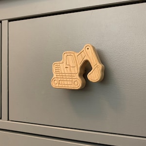 May include: A wooden excavator-shaped cabinet knob. The knob is light brown and attached to a gray cabinet drawer. The excavator design includes details like the cab, arm, and bucket, adding a playful touch to furniture.