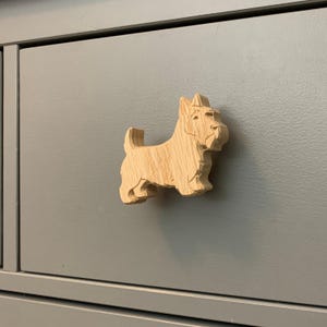 May include: A wooden cabinet knob in the shape of a Scottish Terrier dog. The knob is made of light-colored wood and is attached to a gray cabinet drawer. The dog is carved in detail, showing its distinctive features.