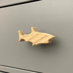 May include: A wooden shark-shaped cabinet knob. The knob is made of light-colored wood and is mounted on a gray cabinet door. The shark design is detailed, with fins and a tail.