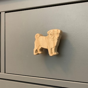 May include: A wooden pug-shaped cabinet knob. The knob is carved from light-coloured wood and features the distinctive features of a pug dog. It is mounted on a grey cabinet drawer, adding a touch of whimsy to furniture.