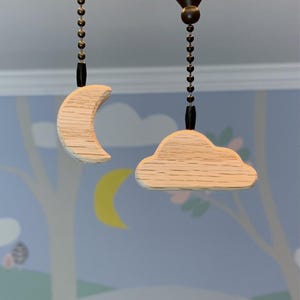 Peut inclure: Deux chaînes de ventilateur de plafond en bois. L'une est en forme de croissant de lune, l'autre en forme de nuage. Les deux sont en bois de couleur claire et sont suspendues à une chaîne sombre.