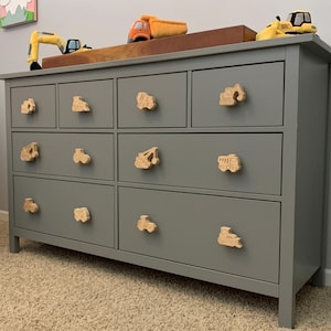 May include: A gray dresser with nine drawers, each featuring a wooden knob shaped like a construction vehicle. The dresser is topped with a wooden shelf holding toy construction vehicles. The knobs are light brown, and the dresser is on a beige carpet.