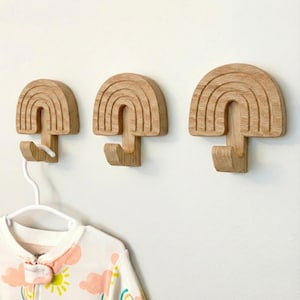 May include: Three wooden rainbow-shaped wall hooks. Each hook is carved from light-colored wood and features a curved rainbow design with a hook extending from the bottom. A white hanger with a baby onesie hangs from one hook.