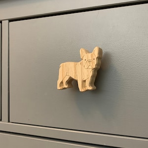 May include: A wooden French Bulldog cabinet knob. The light-coloured wooden knob is attached to a grey cabinet drawer. The dog is depicted standing, with its ears pricked and its tail down. The knob is a decorative furniture accessory.