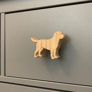 May include: A wooden cabinet knob in the shape of a dog, attached to a gray cabinet drawer. The dog is light brown with visible wood grain and is in a standing position. The cabinet is a solid gray color.