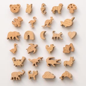 Peut inclure: Une collection de vingt-cinq petites figurines en bois brun clair disposées sur cinq rangées. Les formes incluent des animaux comme un ours, un hibou, un hippocampe, un chien, un flamant rose, un kangourou, un narval, une tortue et un dinosaure.