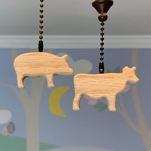 May include: Two wooden fan pull chains, one shaped like a pig and the other shaped like a cow. The chains are made of light brown wood and have a natural finish.