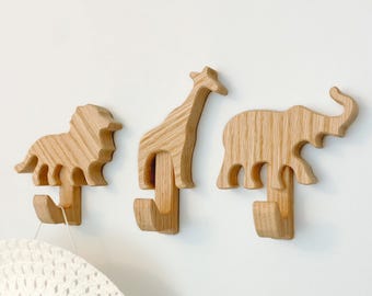 以下が含まれることがあります： ライオン、キリン、ゾウの形をした木製の動物フック3つ。フックは白い壁に取り付けられています。白い帽子がライオンのフックにかけられています。