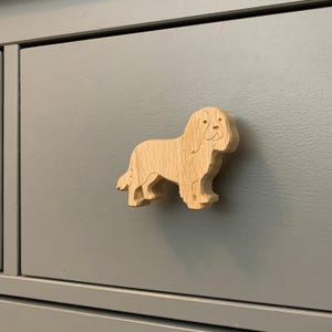 May include: A wooden dog-shaped cabinet knob fixed to a grey drawer. The light brown knob has visible wood grain and a smiling expression. This decorative knob is designed to enhance furniture.