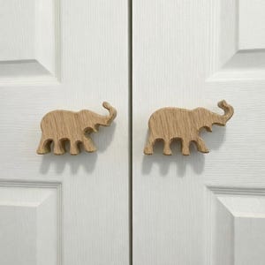 May include: Two wooden elephant-shaped cabinet knobs. The knobs are light brown with visible wood grain and are mounted on a white door. The elephant knobs have a trunk, ears, and four legs.