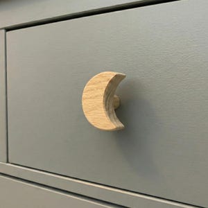 May include: A wooden crescent moon-shaped drawer pull. The drawer pull is made of light-colored wood and is mounted on a gray drawer front. The moon-shaped handle adds a whimsical touch to the furniture.