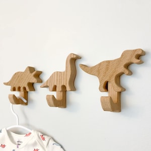 May include: Three wooden dinosaur-shaped hooks mounted on a white wall. The hooks are in the shape of a triceratops, a brachiosaurus, and a tyrannosaurus rex.