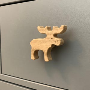 May include: A wooden moose-shaped cabinet knob. The knob is made of light-colored wood and is attached to a gray cabinet door. The moose has antlers, a small eye, and a smiling mouth. The cabinet door is a solid, matte gray.