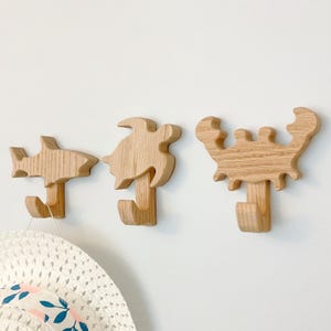 May include: Three wooden wall hooks shaped like a shark, turtle, and crab. The hooks are painted in a natural wood color. A white hat is hanging on the shark hook.
