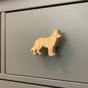 May include: A wooden cabinet knob in the shape of a German Shepherd dog. The knob is made of light-colored wood and is attached to a gray cabinet drawer. The dog is depicted in a standing pose.