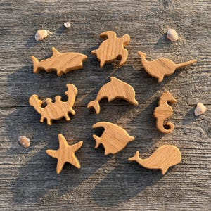 May include: Wooden sea creature cutouts including a shark, turtle, dolphin, seahorse, crab, starfish, fish, and whale. The wooden figures are light brown and arranged on a weathered wooden surface with seashells.