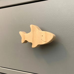 May include: A wooden shark-shaped cabinet knob with a smiling face is attached to a grey cabinet. The knob is made of light-coloured wood and has a smooth finish. The cabinet has a simple, modern design.