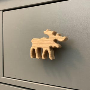 Peut inclure: Bouton de meuble en forme d'élan en bois clair. Le bouton est monté sur un tiroir de meuble gris. Le design de l'élan présente des bois et quatre pattes, ajoutant une touche rustique au meuble. Mesure environ 7 cm de long.