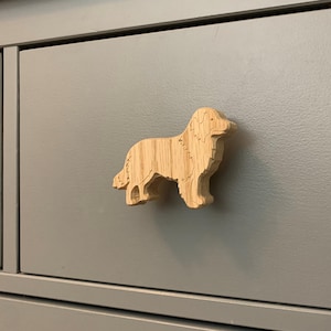 May include: A wooden dog-shaped cabinet knob, possibly a Golden Retriever, attached to a grey drawer. The knob is crafted from light-coloured wood, featuring a detailed, layered design, suitable for a cabinet or drawer.
