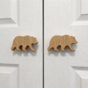 May include: Two wooden bear-shaped cabinet pulls. The pulls are made of light-coloured wood and are attached to a white door. The bears are in a walking pose, with their legs and bodies carved in detail.