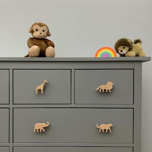 Peut inclure: Une commode grise à six tiroirs avec des poignées de tiroir en bois en forme d'animaux. Les poignées comprennent une girafe, un lion, un éléphant et un tigre. Un jouet arc-en-ciel et un lion en peluche sont posés sur la commode.
