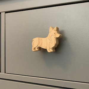 May include: A wooden corgi-shaped drawer pull. The pull is made of light-colored wood and is attached to a gray drawer. The corgi is depicted in a standing position, with its ears perked up.
