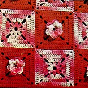 May include: A crocheted blanket with a square pattern in shades of red, pink, and white. The blanket features a floral design with a central flower in each square.