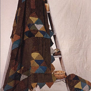 Puede incluir: Una manta de punto marrón con un patrón geométrico de triángulos en tonos marrones, verdes, azules y grises. La manta está drapeada sobre una escalera de madera y una almohada más pequeña con el mismo patrón está en el suelo.
