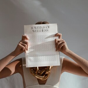 May include: A person is holding a white sheet of paper in front of their face. The paper has the text "8 Weeks of Wellness" printed on it. The paper is divided into eight sections, each with a grid of squares. The person is wearing a white knitted top and a gold chain necklace.