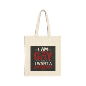 May include: Cream-colored tote bag with a black and red graphic. The graphic reads "I AM GAY and I WANT A HUSBAND" in a bold, stylized font. The bag has a simple design with two handles for carrying.