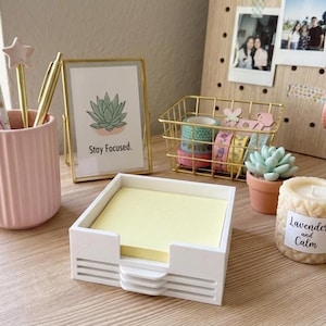 May include: A white square notepad holder with yellow notepaper sits on a wooden desk. A gold-framed print with a succulent illustration and the text "Stay Focused" is nearby. Other items include a pink pen holder, decorative tape, and a candle.