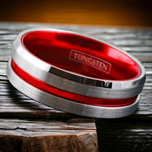 May include: A silver tungsten ring with a red inlay and the word "TUNGSTEN" engraved on the inside of the ring.